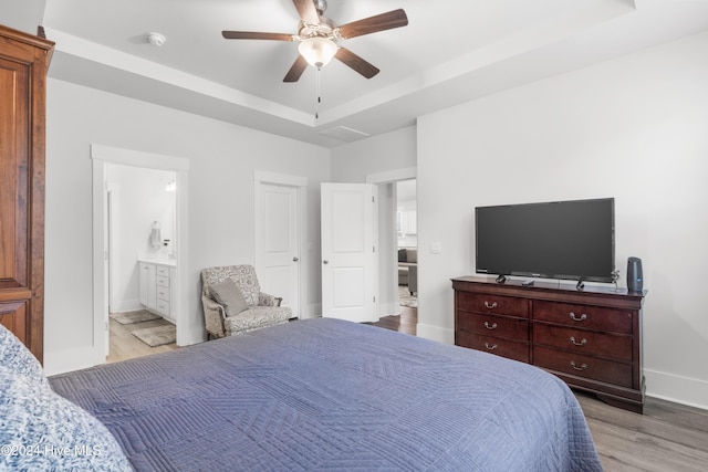 bedroom with a tray ceiling, ensuite bath, ceiling fan, and light wood-type flooring