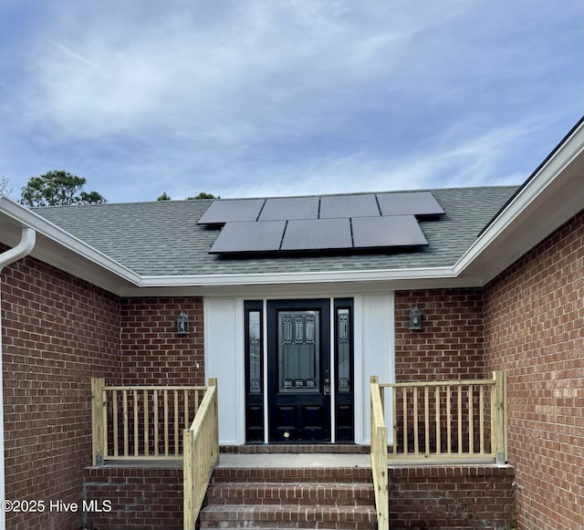 doorway to property featuring a shingled roof, brick siding, and roof mounted solar panels