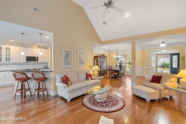 living room with ornate columns, light hardwood / wood-style flooring, high vaulted ceiling, ceiling fan with notable chandelier, and ornamental molding