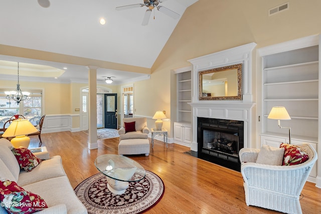 living room featuring light wood-type flooring, ornate columns, ornamental molding, ceiling fan with notable chandelier, and built in features