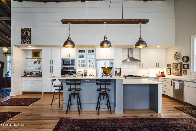 kitchen featuring wall chimney exhaust hood, stainless steel appliances, a towering ceiling, decorative light fixtures, and white cabinets