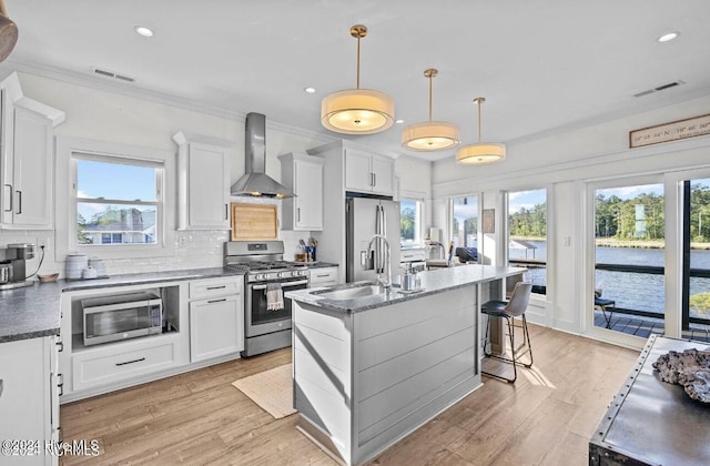 kitchen with white cabinetry, wall chimney exhaust hood, hanging light fixtures, an island with sink, and appliances with stainless steel finishes