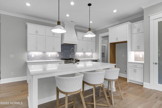 kitchen featuring custom range hood, a center island with sink, light hardwood / wood-style flooring, white cabinets, and hanging light fixtures