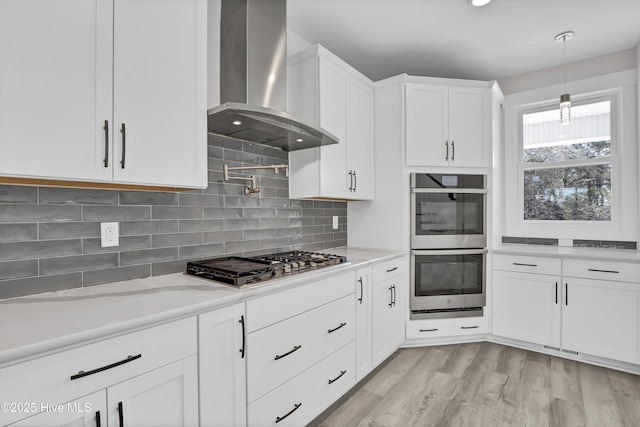 kitchen featuring wall chimney range hood, stainless steel appliances, white cabinets, decorative light fixtures, and light wood-type flooring