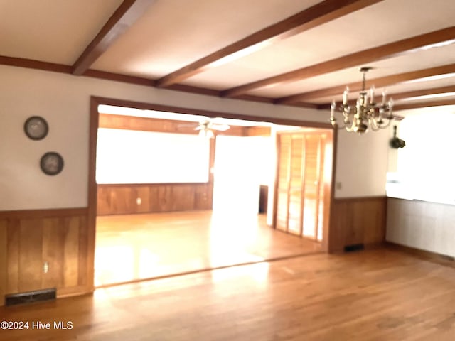 empty room featuring ceiling fan with notable chandelier, beam ceiling, hardwood / wood-style floors, and wooden walls