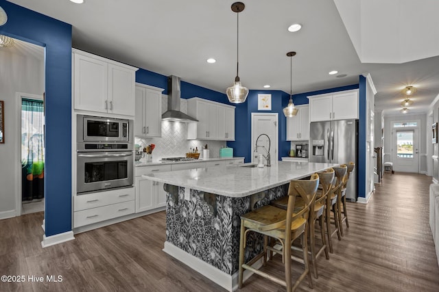 kitchen with decorative backsplash, appliances with stainless steel finishes, wall chimney exhaust hood, white cabinets, and an island with sink