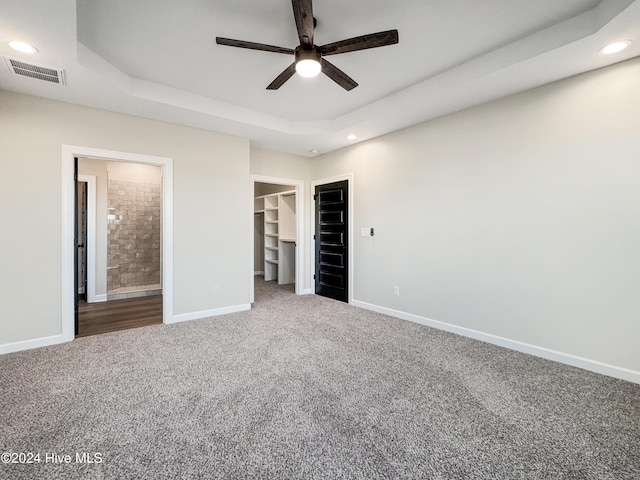 unfurnished bedroom featuring a walk in closet, a tray ceiling, ensuite bath, and ceiling fan