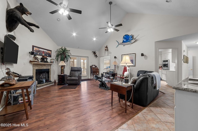 living area with a fireplace with raised hearth, ceiling fan, light wood-type flooring, french doors, and high vaulted ceiling