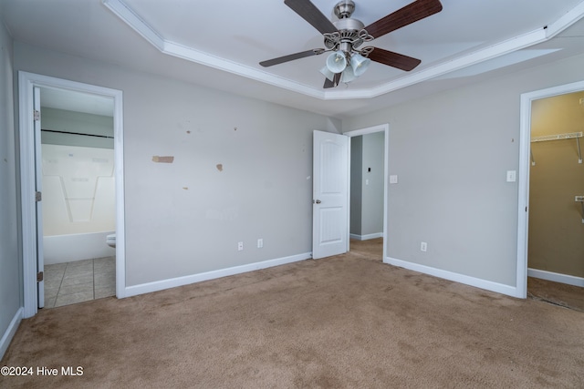 unfurnished bedroom with light carpet, ceiling fan, a spacious closet, connected bathroom, and a tray ceiling