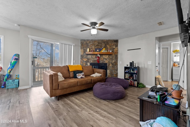 living room featuring ceiling fan, a stone fireplace, light wood-type flooring, and a textured ceiling