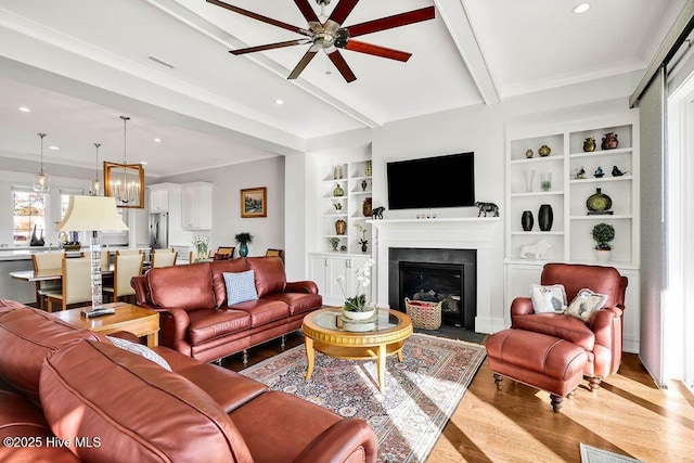 living room featuring built in features, beamed ceiling, ceiling fan with notable chandelier, and light wood-type flooring