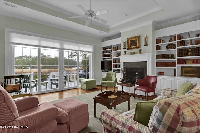 living room featuring built in features, a high end fireplace, a tray ceiling, crown molding, and light hardwood / wood-style flooring