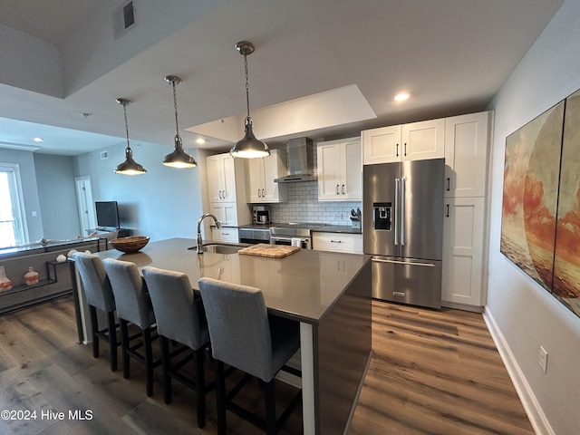 kitchen with white cabinetry, a center island with sink, hanging light fixtures, appliances with stainless steel finishes, and wall chimney range hood