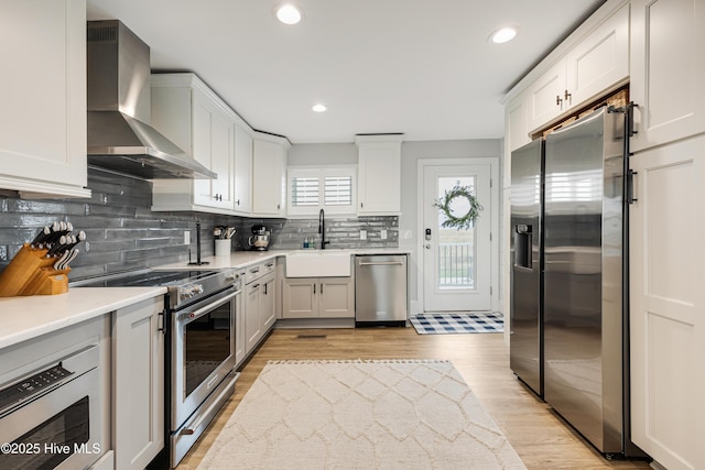 kitchen with light wood-style floors, appliances with stainless steel finishes, light countertops, wall chimney range hood, and a sink