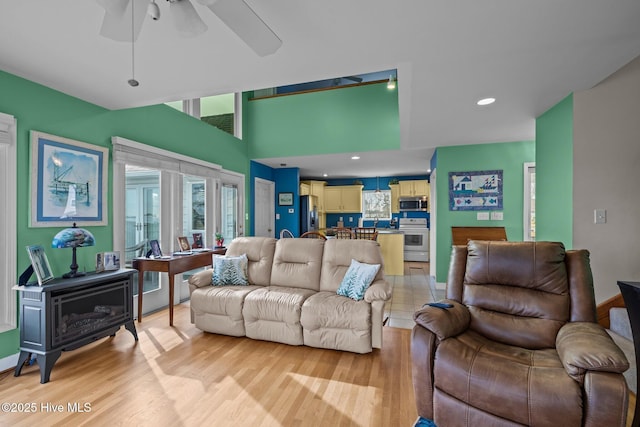 living room featuring ceiling fan, light wood-type flooring, a wood stove, and a towering ceiling