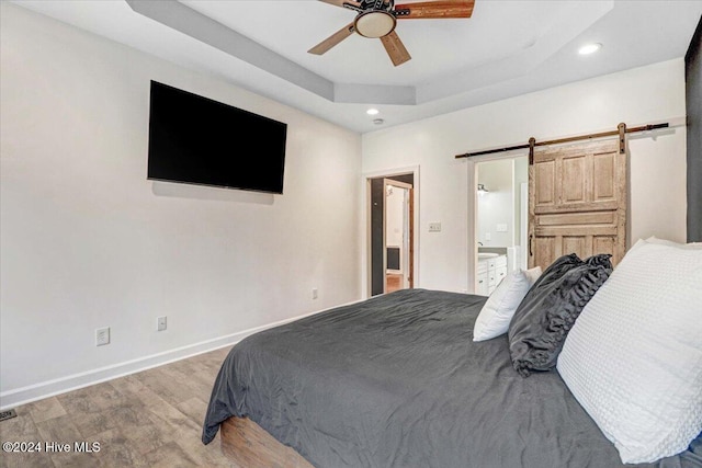 bedroom featuring a tray ceiling, ceiling fan, a barn door, light hardwood / wood-style flooring, and connected bathroom