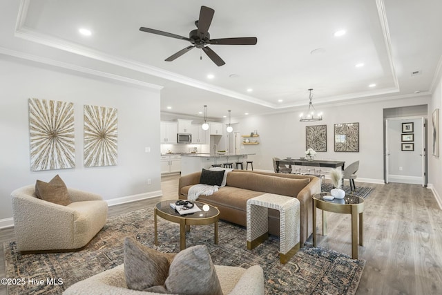 living area with a tray ceiling, baseboards, light wood-type flooring, and ceiling fan with notable chandelier