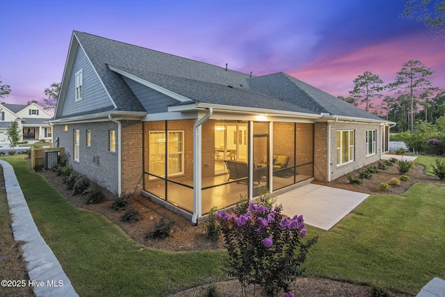 back of property at dusk featuring a sunroom, a shingled roof, brick siding, and a yard