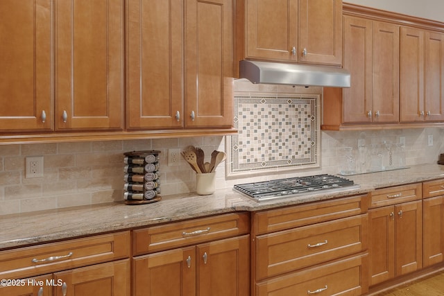 kitchen featuring decorative backsplash, brown cabinetry, light stone countertops, under cabinet range hood, and stainless steel gas cooktop