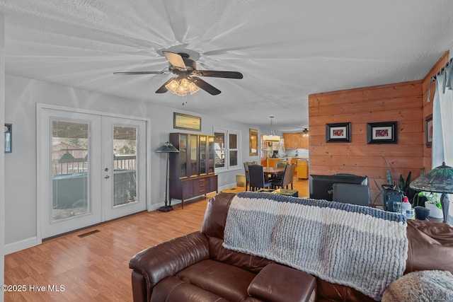 living room with ceiling fan, french doors, light hardwood / wood-style flooring, wood walls, and a textured ceiling