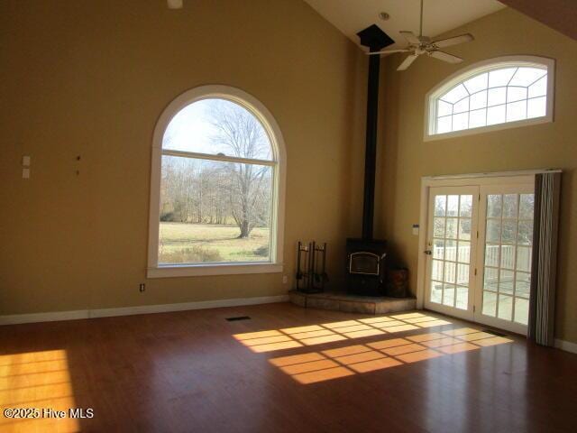 unfurnished living room with a wood stove, ceiling fan, wood finished floors, high vaulted ceiling, and baseboards