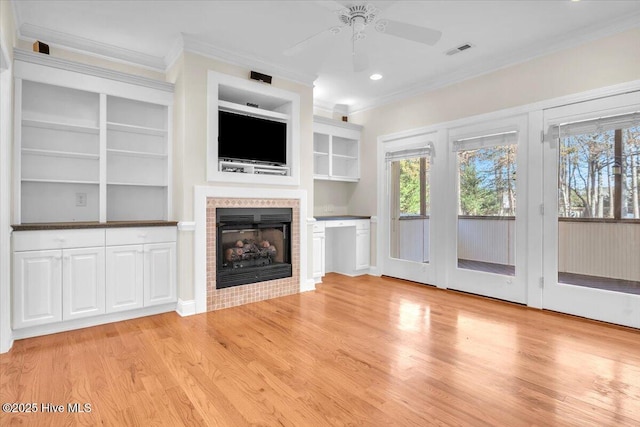 unfurnished living room featuring a tile fireplace, ceiling fan, built in shelves, crown molding, and light wood-type flooring