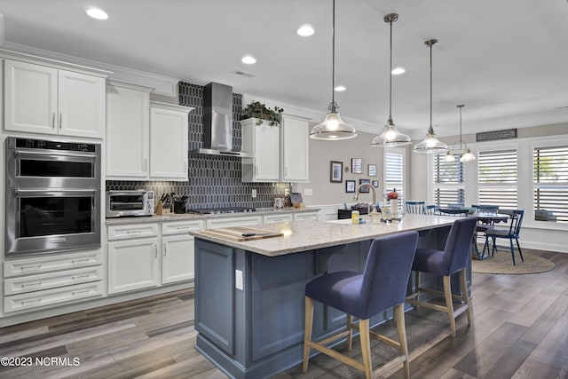 kitchen featuring white cabinetry, stainless steel double oven, hanging light fixtures, wall chimney range hood, and a kitchen island with sink