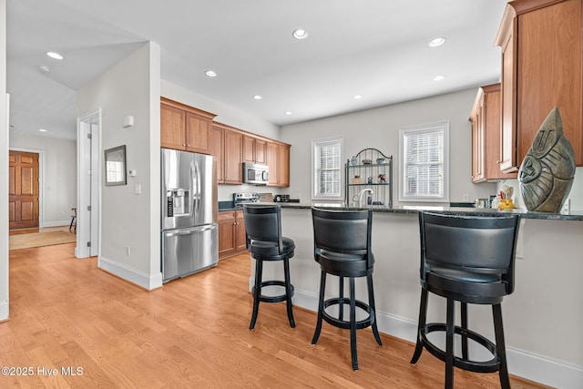 kitchen with kitchen peninsula, appliances with stainless steel finishes, dark stone counters, light hardwood / wood-style flooring, and a breakfast bar