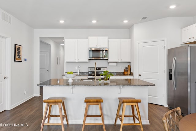 kitchen with white cabinetry, appliances with stainless steel finishes, a kitchen island with sink, dark wood-type flooring, and dark stone countertops