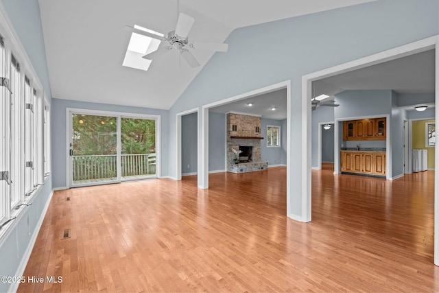 unfurnished living room featuring high vaulted ceiling, a skylight, a brick fireplace, ceiling fan, and light wood-type flooring