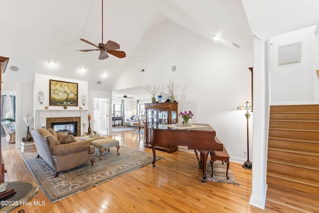 living area with a fireplace, light wood-style flooring, a ceiling fan, high vaulted ceiling, and stairs