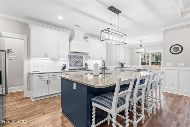 kitchen featuring premium range hood, ornamental molding, stainless steel gas stovetop, white cabinetry, and a kitchen island with sink