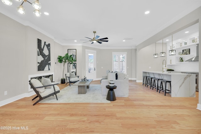 living room with ceiling fan with notable chandelier, ornamental molding, sink, and light hardwood / wood-style floors