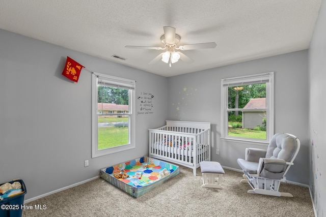 carpeted bedroom with a textured ceiling, multiple windows, ceiling fan, and a crib