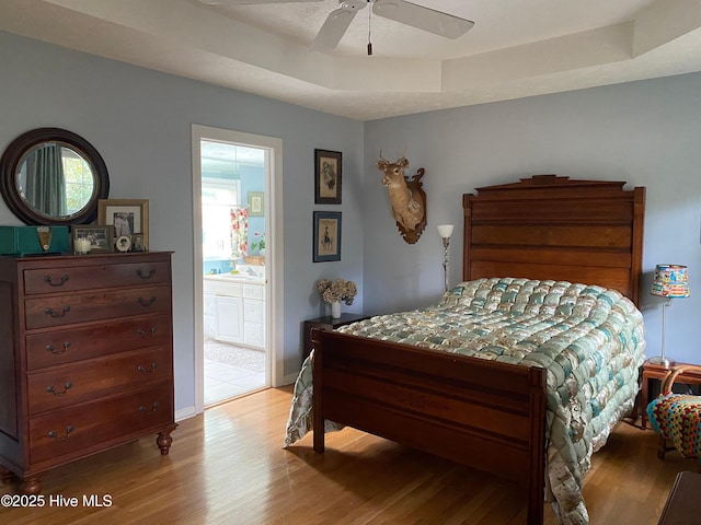 bedroom featuring a tray ceiling, ensuite bathroom, ceiling fan, and light wood-type flooring