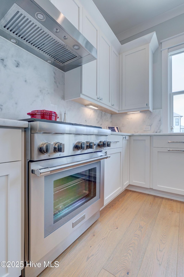 kitchen featuring wall chimney range hood, tasteful backsplash, high end stove, white cabinets, and light wood-type flooring