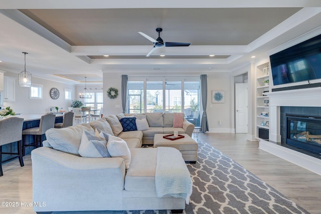 living room with a tile fireplace, built in shelves, light wood-type flooring, ceiling fan with notable chandelier, and ornamental molding