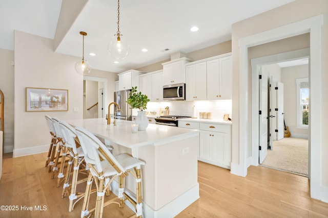 kitchen with pendant lighting, stainless steel appliances, light wood-type flooring, a kitchen island with sink, and white cabinetry