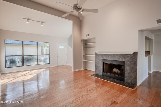 unfurnished living room featuring ceiling fan, wood-type flooring, a tiled fireplace, and built in shelves