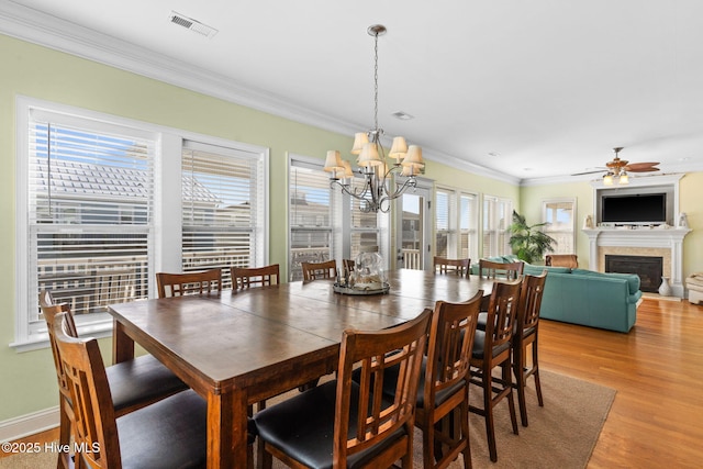 dining space with crown molding, a glass covered fireplace, ceiling fan with notable chandelier, light wood-type flooring, and visible vents