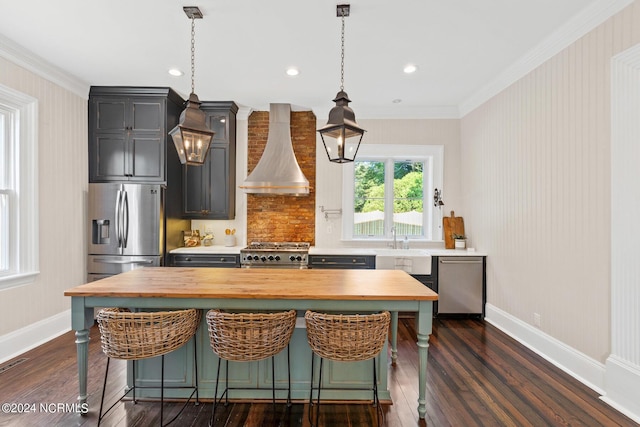 kitchen featuring appliances with stainless steel finishes, wall chimney exhaust hood, decorative light fixtures, a kitchen breakfast bar, and butcher block countertops