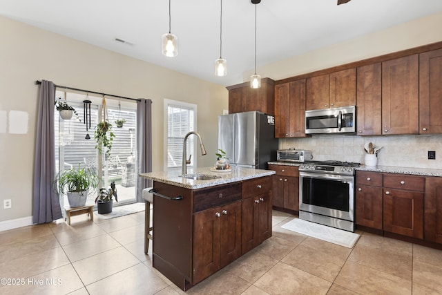 kitchen featuring appliances with stainless steel finishes, tasteful backsplash, sink, light stone counters, and a center island with sink