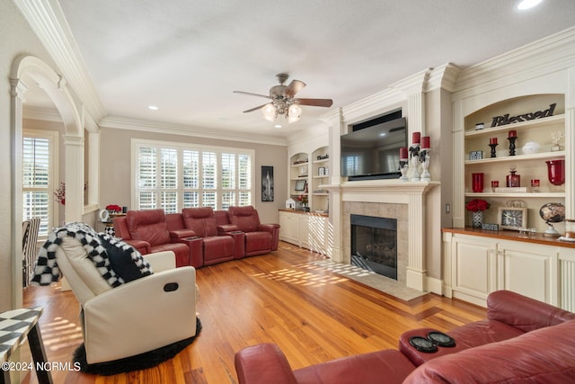 living room with built in features, a tile fireplace, ceiling fan, ornamental molding, and light wood-type flooring