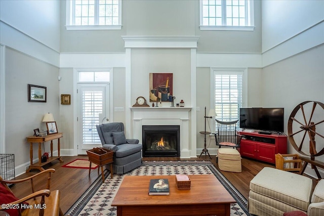 living area featuring a high ceiling, a lit fireplace, a healthy amount of sunlight, and dark wood-style flooring