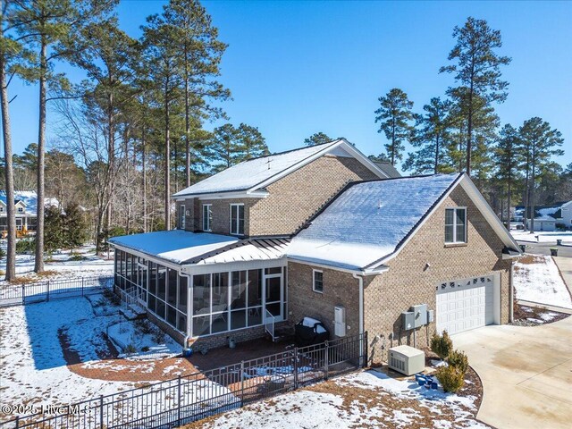 snow covered back of property featuring fence, concrete driveway, a sunroom, a garage, and brick siding