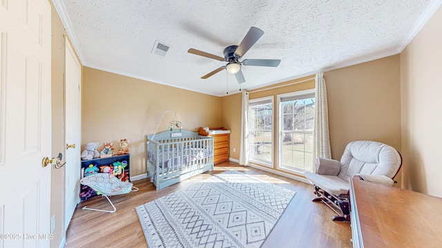 bedroom with ceiling fan, light hardwood / wood-style flooring, a textured ceiling, a crib, and ornamental molding