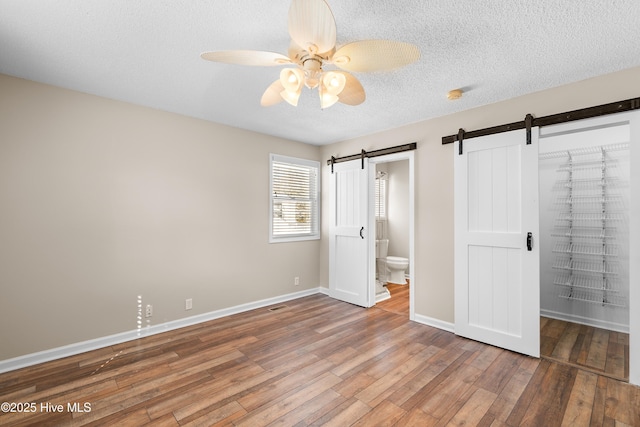 unfurnished bedroom with ensuite bathroom, a barn door, ceiling fan, hardwood / wood-style flooring, and a textured ceiling