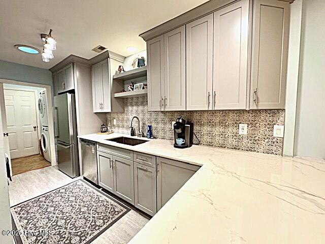 kitchen featuring sink, stacked washing maching and dryer, light stone countertops, appliances with stainless steel finishes, and gray cabinetry