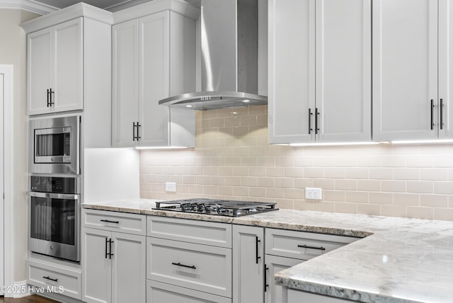 kitchen with white cabinets, wall chimney range hood, decorative backsplash, light stone countertops, and stainless steel appliances