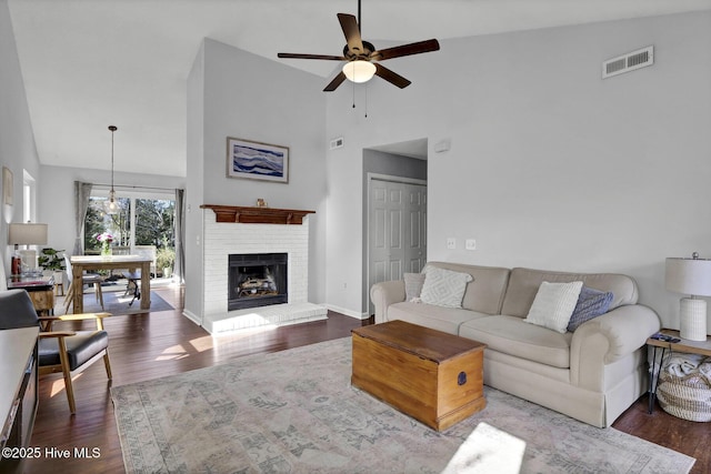 living room featuring dark wood-type flooring, a towering ceiling, ceiling fan, and a brick fireplace